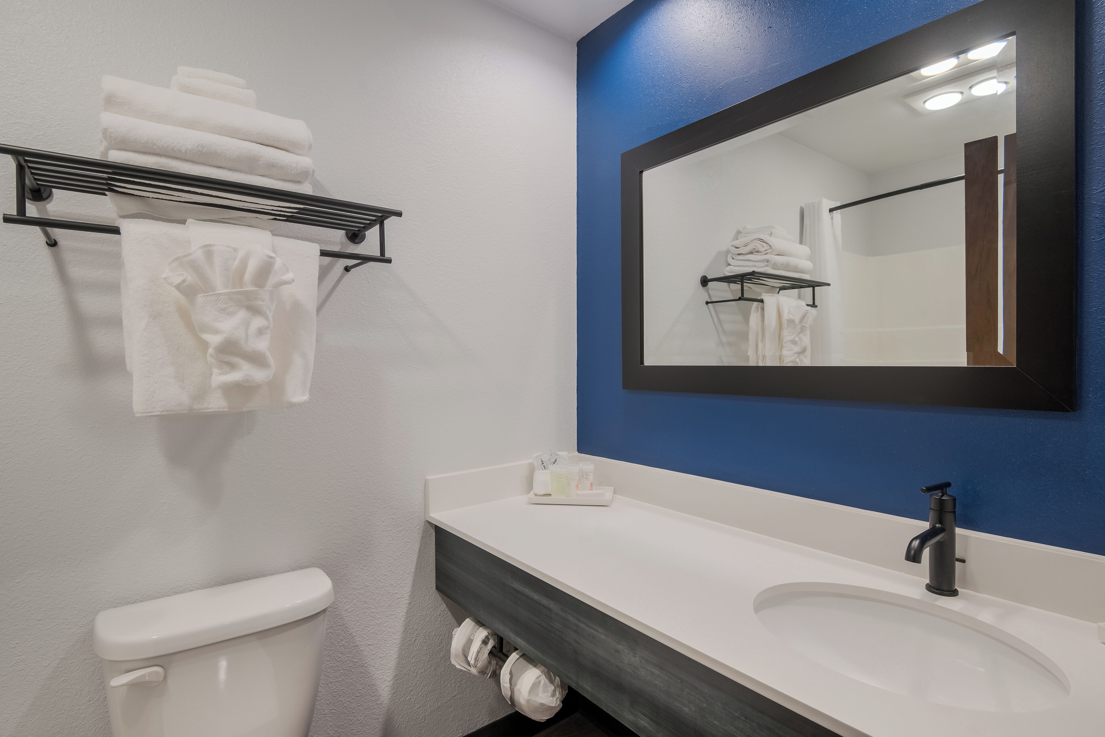 A bathroom with a white toilet, a white sink, a towel rack with folded towels, and a mirror with a blue accent wall.