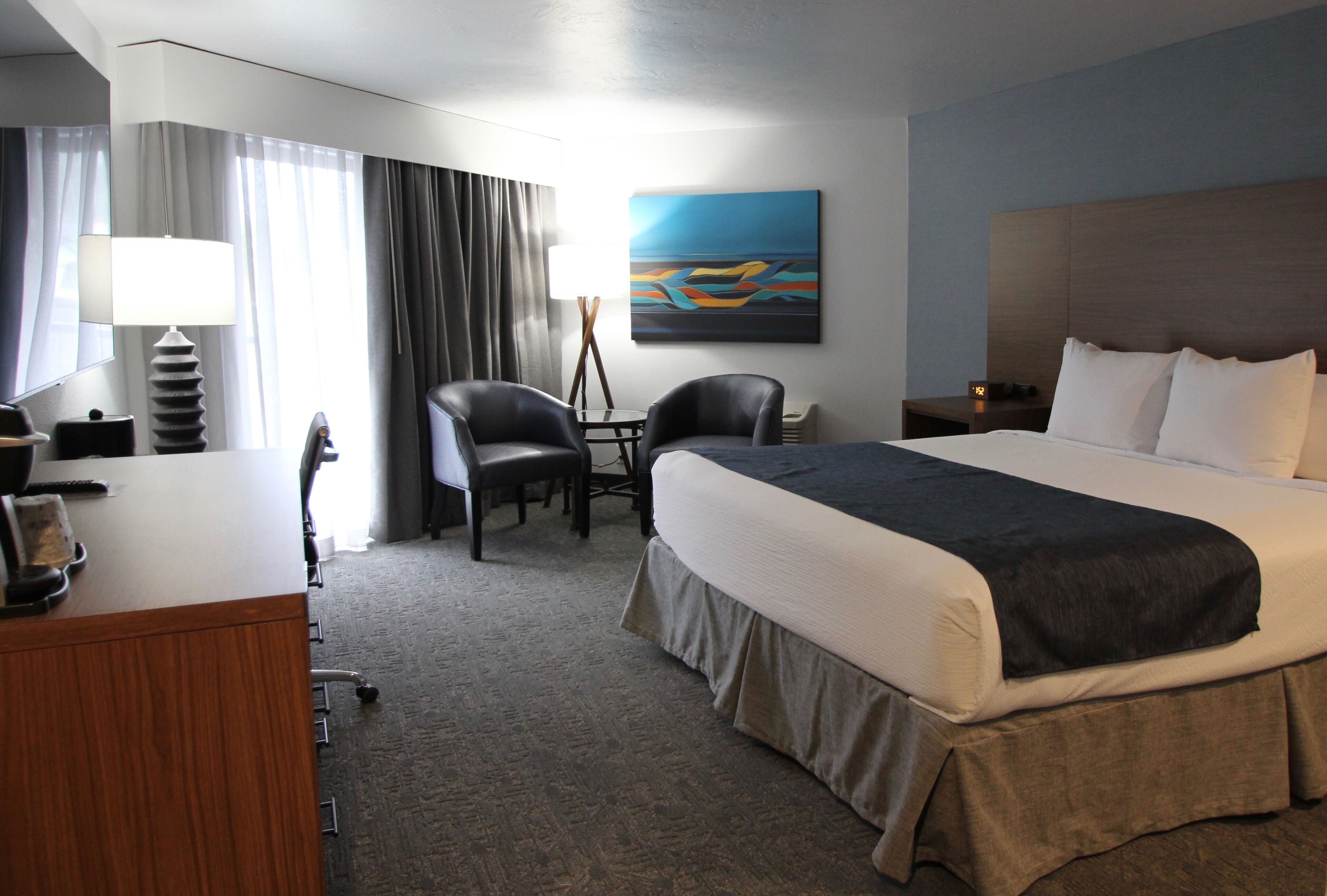 A hotel room with a large bed, a desk with a lamp, a chair, a small table, and a painting on the wall.