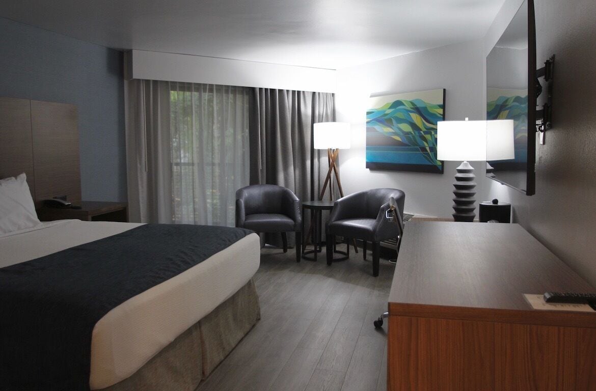 A hotel room with a bed, two chairs, a desk, a lamp, and a painting on the wall.
