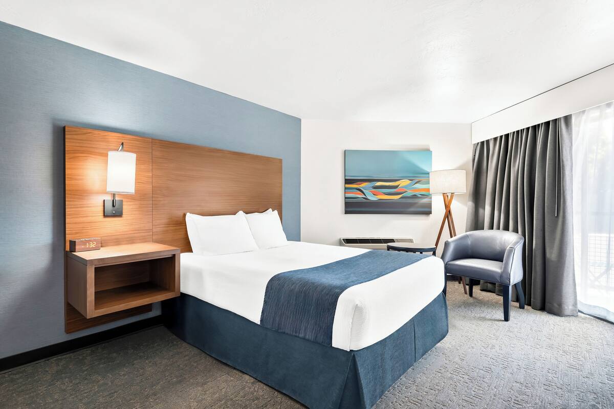 A hotel room with a bed, a nightstand, a desk, a chair, a painting, and a window with curtains.