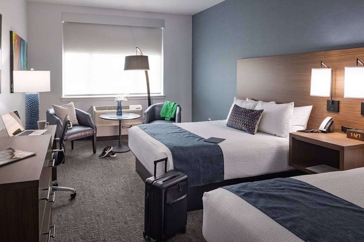 A hotel room with two beds, a desk with a laptop, a chair, a small table with a lamp, and a suitcase.