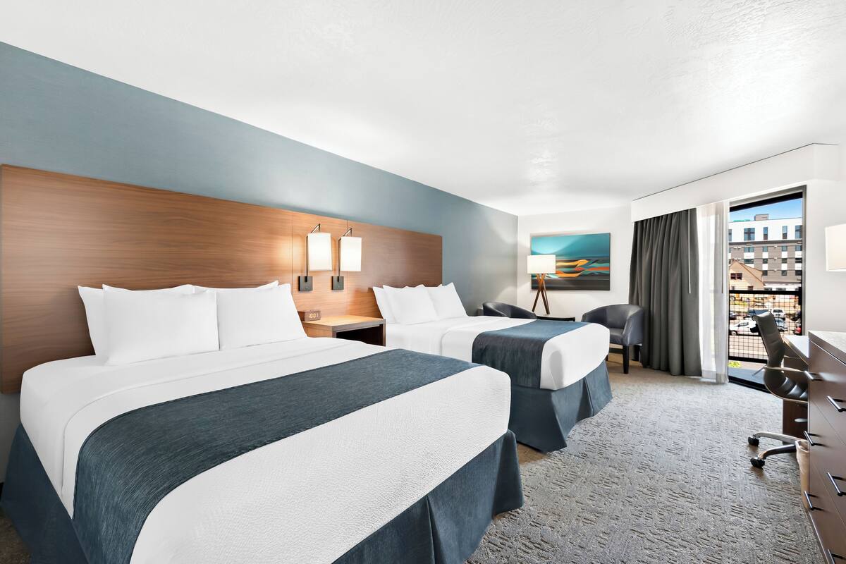A hotel room with two beds, a desk, a chair, and a large window with a view of buildings.