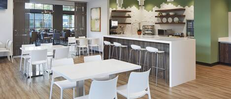 A modern dining area with white tables and chairs, a central bar area with stools, and large windows providing natural light.