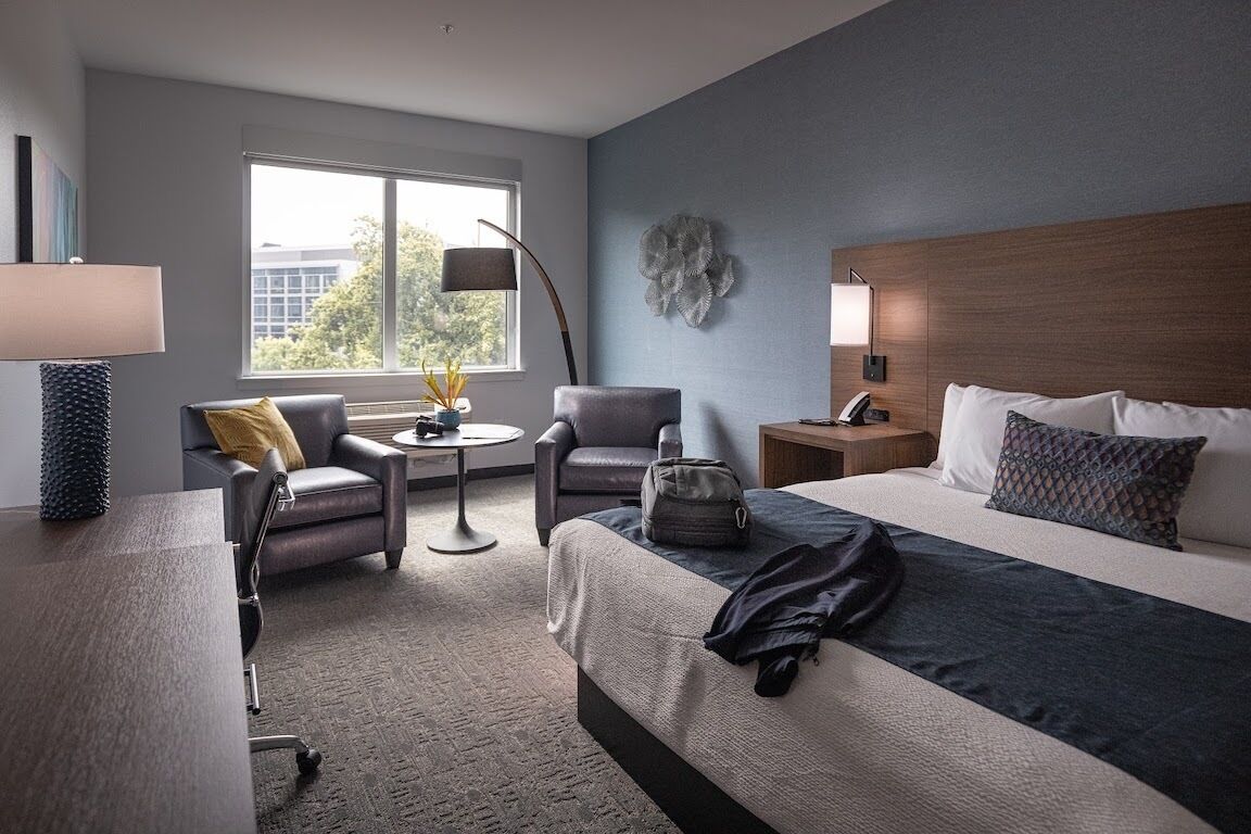 Habitación de hotel con cama, dos sillones, una mesita redonda, lámpara, mesita de noche y ventana con vistas a edificios.