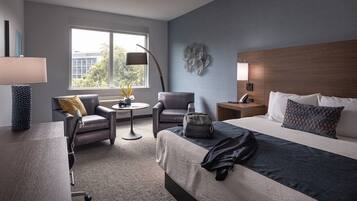 Habitación de hotel con cama, dos sillones, una mesita redonda, lámpara, mesita de noche y ventana con vistas a edificios.