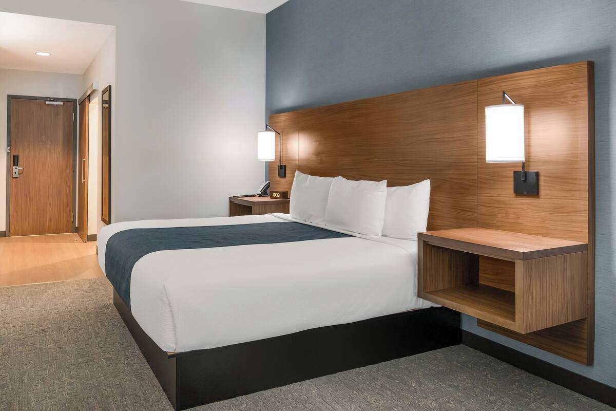 A modern hotel room with a large bed, wooden headboard, bedside tables, and wall-mounted lamps.