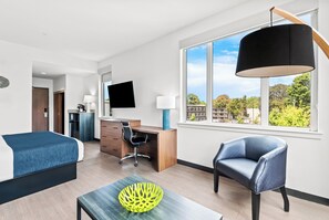 A modern hotel room with a bed, desk, chair, and a large window with a city view.