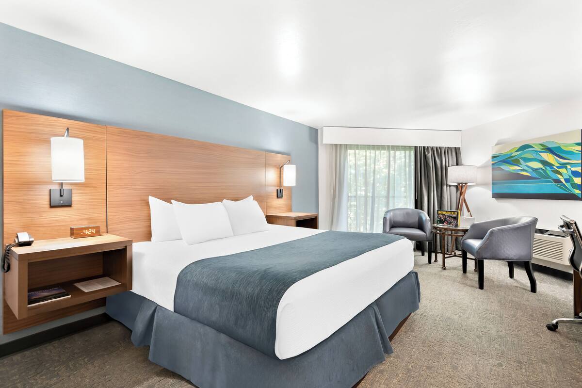 A hotel room with a large bed, bedside tables, a desk, a chair, and a painting on the wall.