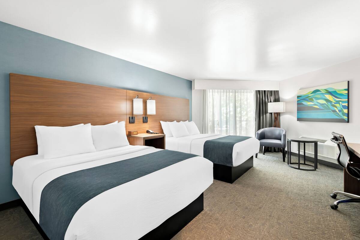 A hotel room with two beds, a desk, a chair, a lamp, and a painting on the wall.