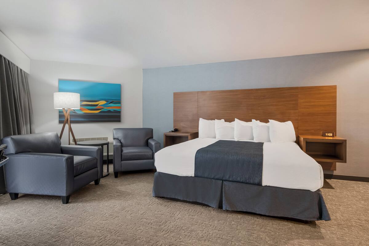 A hotel room with a large bed, two armchairs, a side table, a lamp, a painting on the wall, and a wooden headboard.