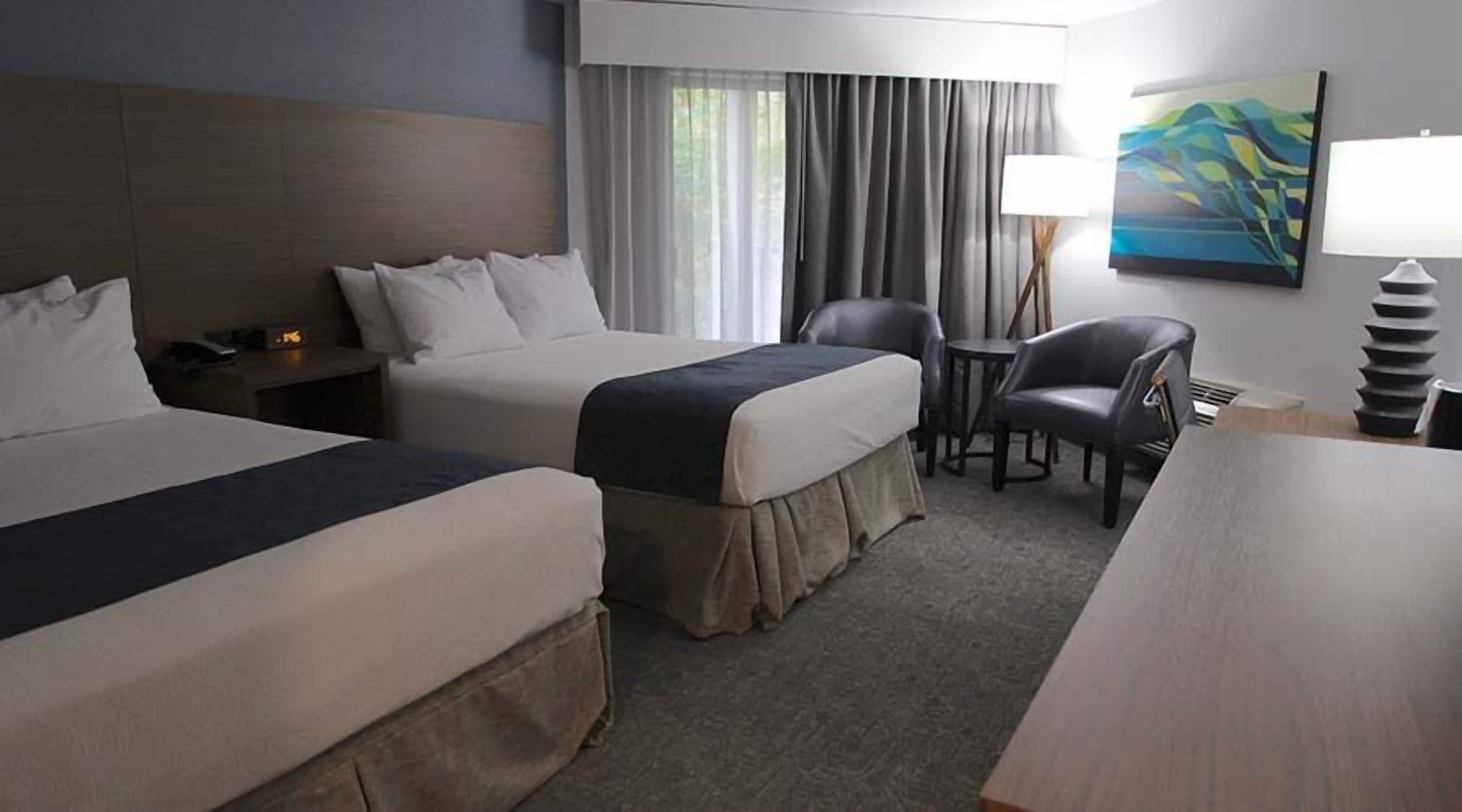 Habitación de hotel con dos camas, un escritorio, una silla, una lámpara y un cuadro en la pared.