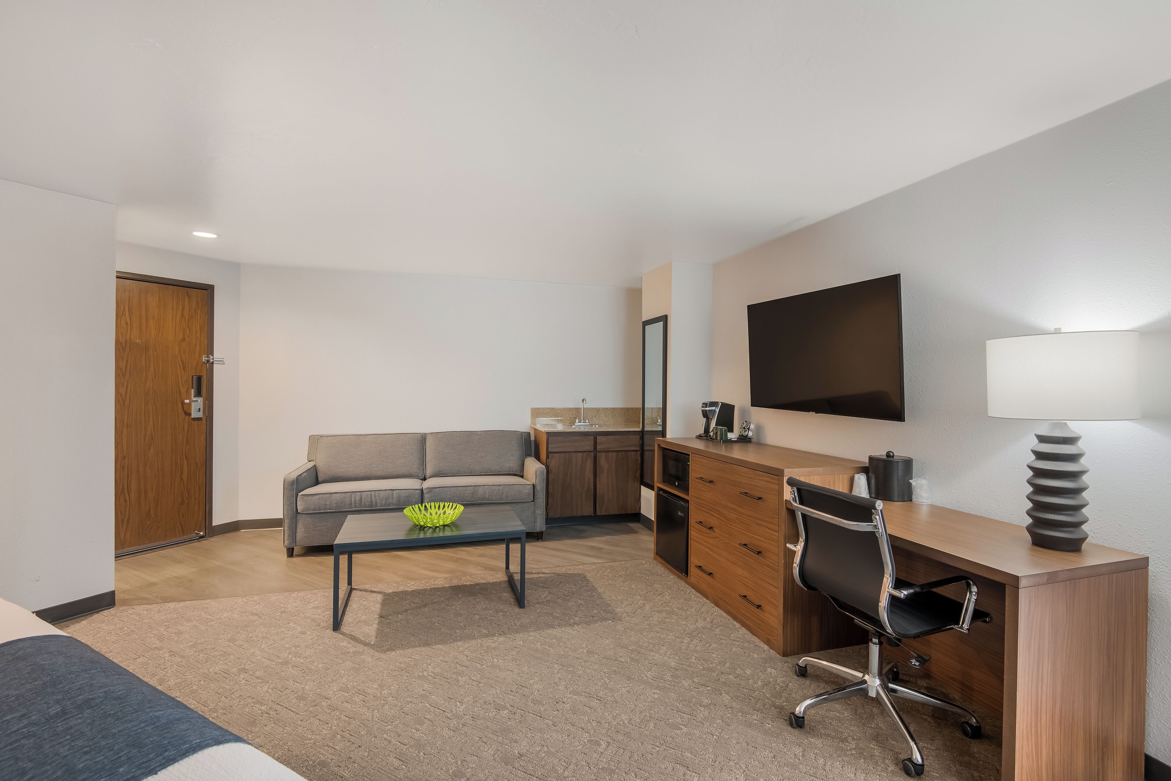 A modern hotel room with a sofa, a desk, a chair, a coffee table, a TV, and a kitchen area.