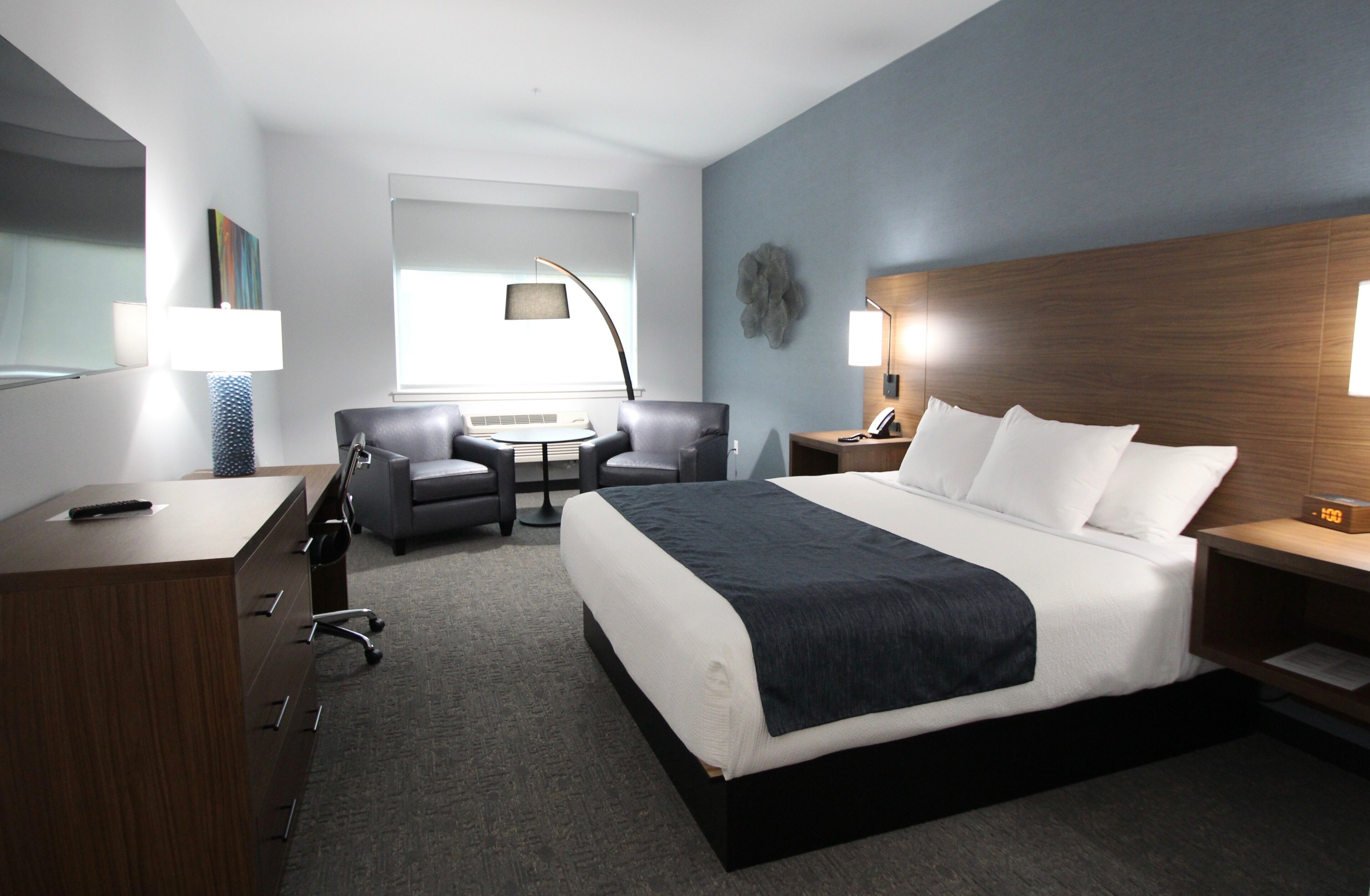 A modern hotel room with a large bed, a desk with drawers, a chair, a small table, and a lamp.
