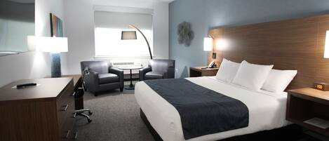 A modern hotel room with a large bed, a desk with drawers, a chair, a small table, and a lamp.