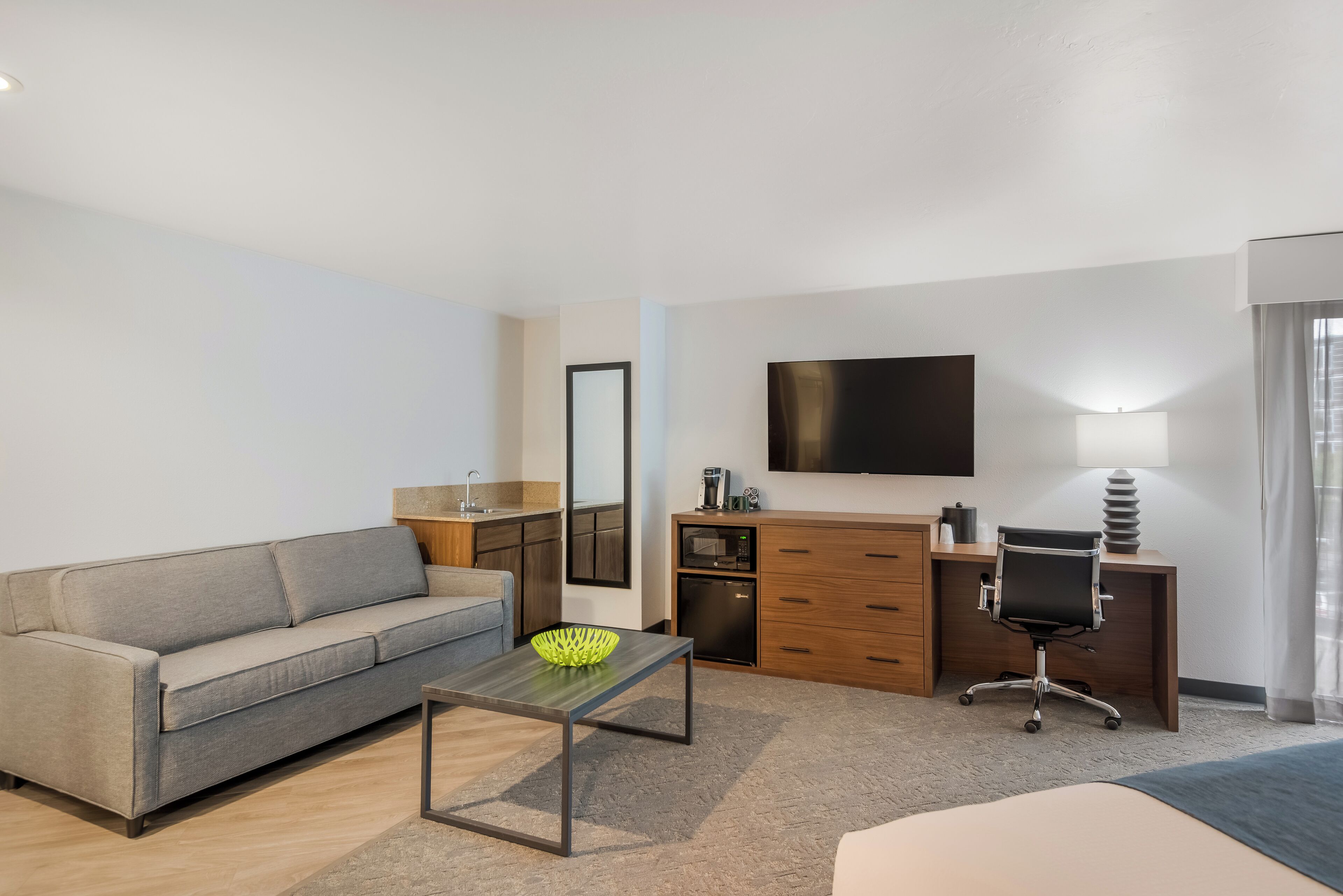 Une chambre d’hôtel moderne équipée d’un canapé, d’une table basse, d’un bureau avec une chaise, d’un téléviseur à écran plat, d’un miroir et d’une kitchenette.