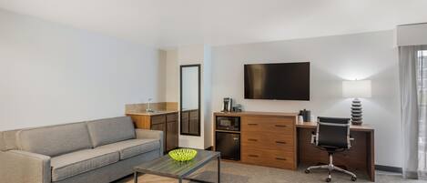 Una habitación de hotel moderna con sofá, una mesa de centro, un escritorio con silla, un televisor de pantalla plana, un espejo y una kitchenette.