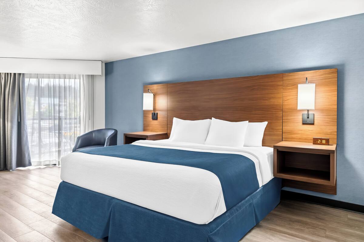 A hotel room with a large bed, wooden headboard, two bedside tables with lamps, a chair, and a view of the outdoors through the window.