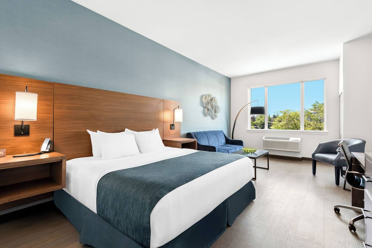 A modern hotel room with a large bed, a desk, a chair, and a window with a view of trees.