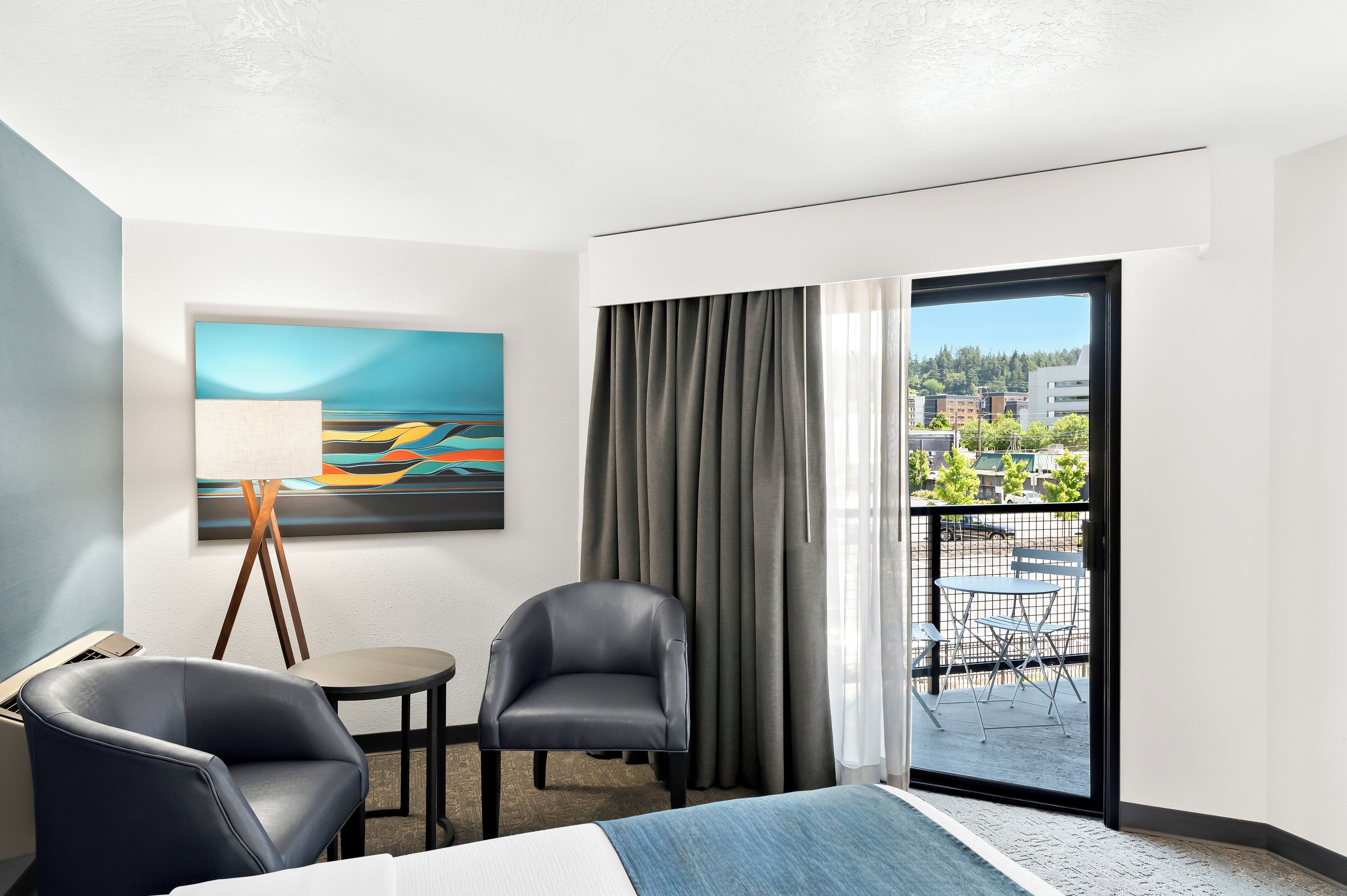 Habitación de hotel con cama, dos sillas, una mesita, un cuadro en la pared y vistas al exterior por una puerta corredera.