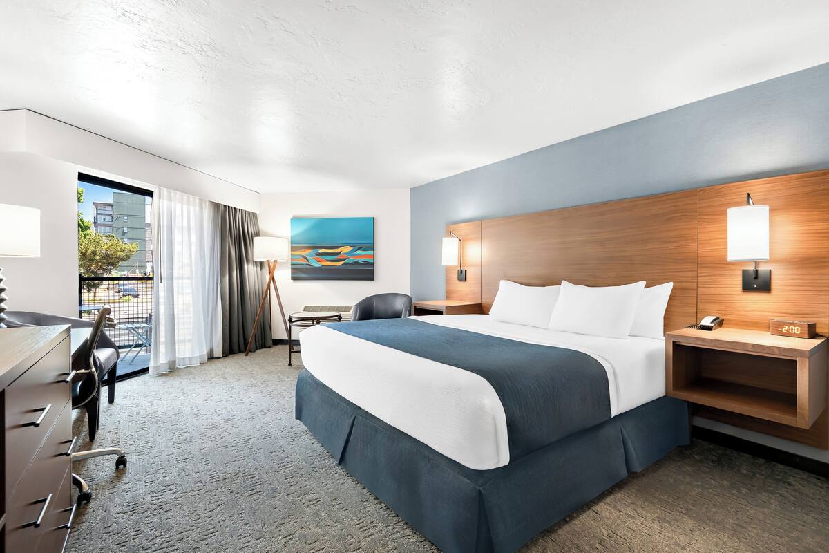 A hotel room with a large bed, a desk with a chair, a nightstand with a lamp, and a painting on the wall.
