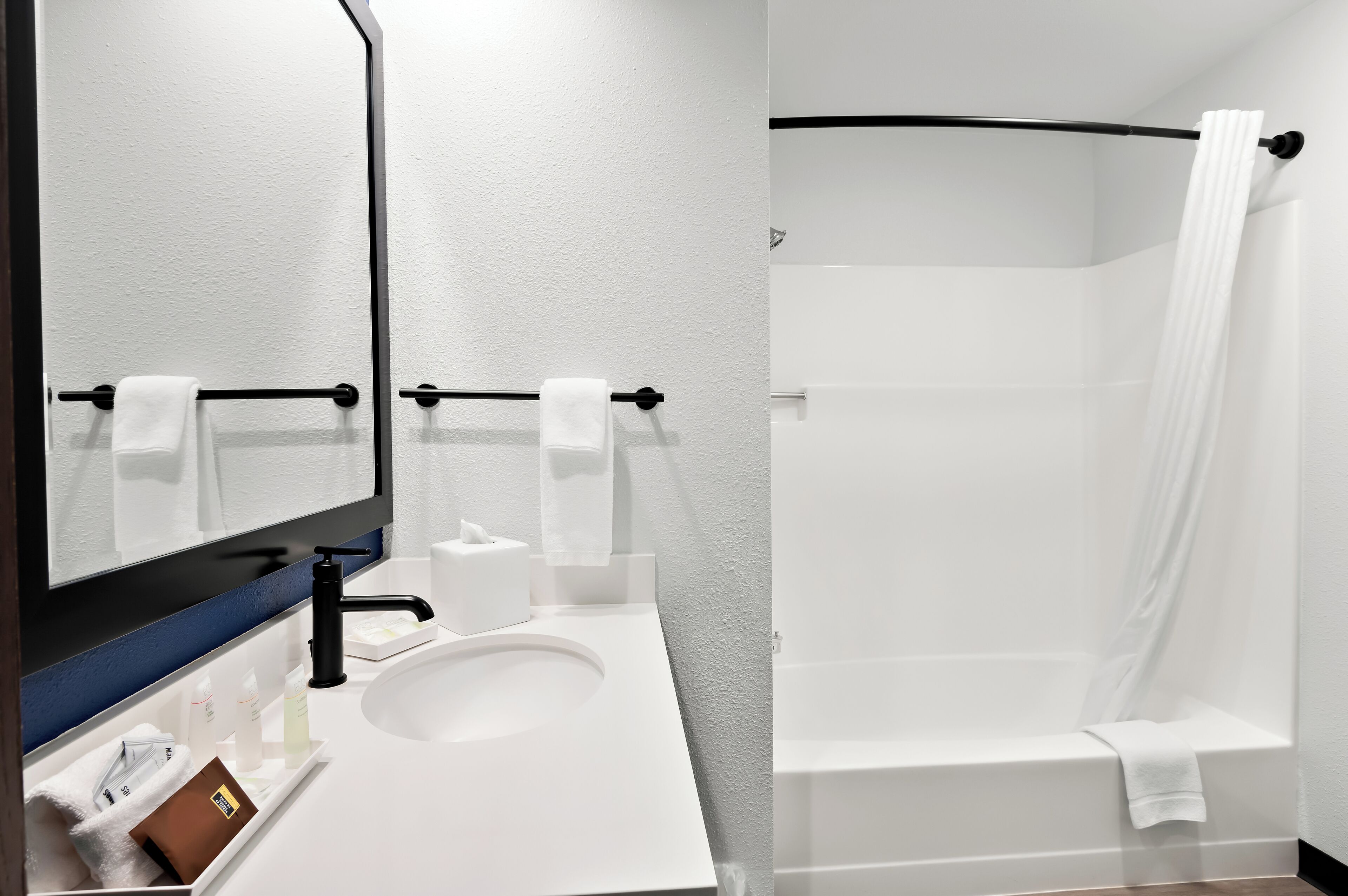Une salle de bain moderne avec un lavabo blanc, un robinet noir et un grand miroir. On y trouve un porte-serviettes avec des serviettes, une baignoire blanche et un rideau de douche.
