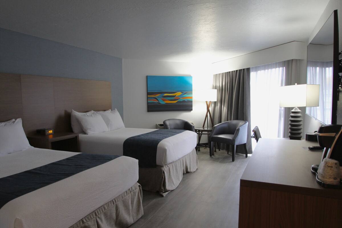 A hotel room with two beds, a desk, a chair, a lamp, and a painting on the wall.