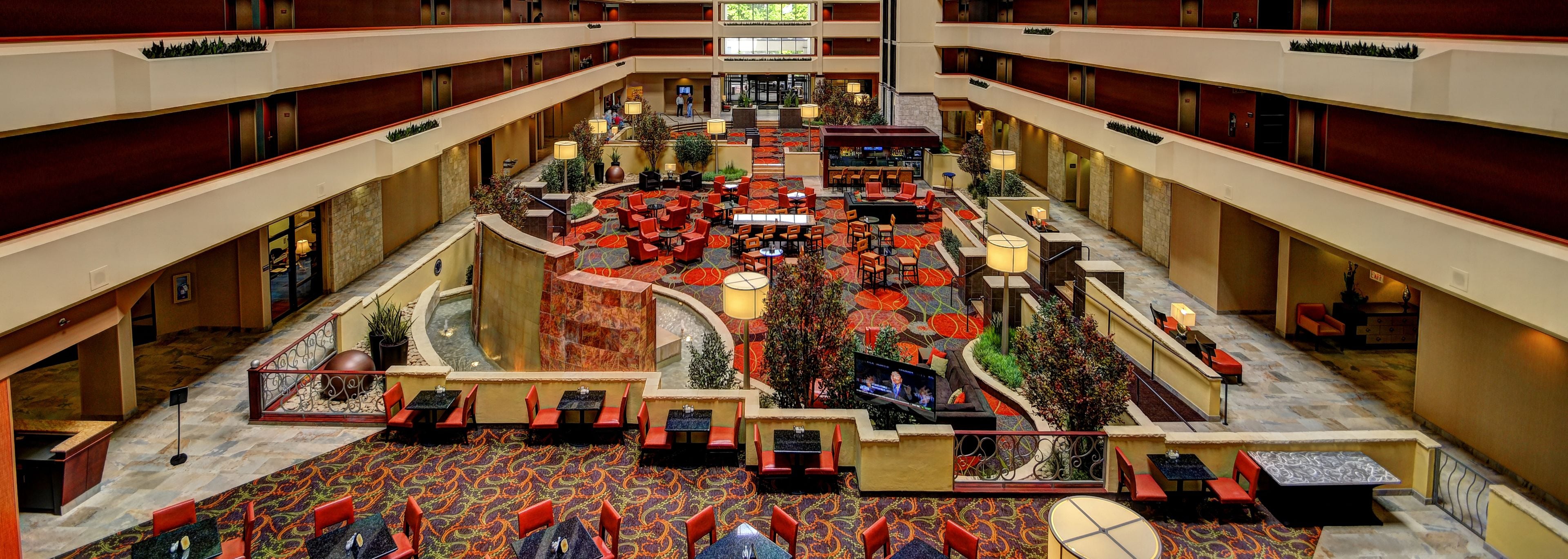 Hall d'ingresso di un hotel su più livelli, con un'ampia atrio centrale, diverse zone relax e uno schermo televisivo.