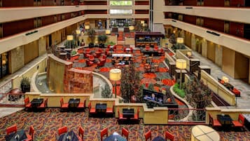 Hall d'ingresso di un hotel su più livelli, con un'ampia atrio centrale, diverse zone relax e uno schermo televisivo.
