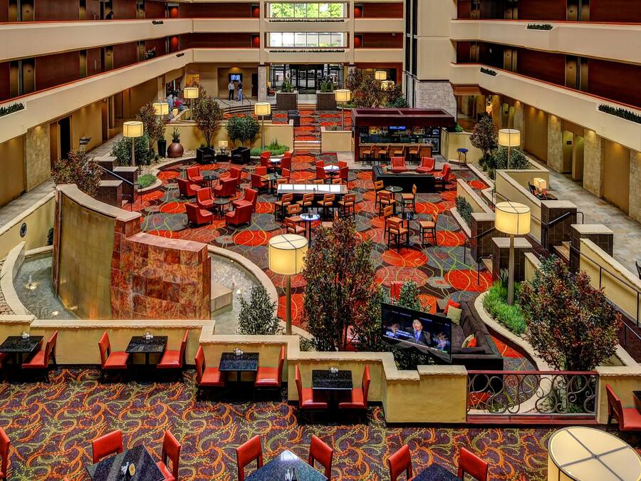 Hall d'ingresso di un hotel su più livelli, con un'ampia atrio centrale, diverse zone relax e uno schermo televisivo.