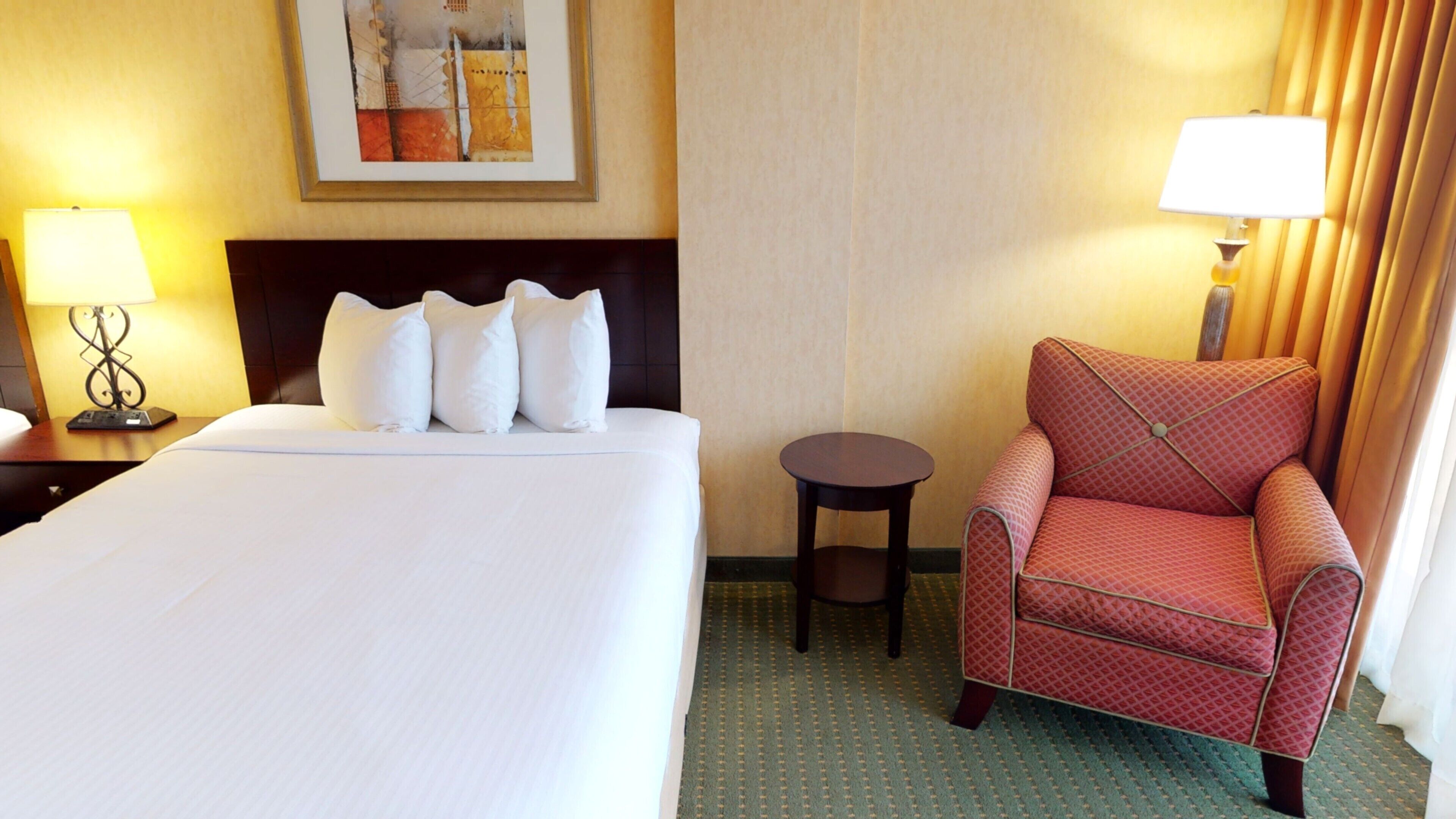 Une chambre d’hôtel avec un grand lit, un fauteuil rouge à motifs, une petite table ronde et deux lampes.