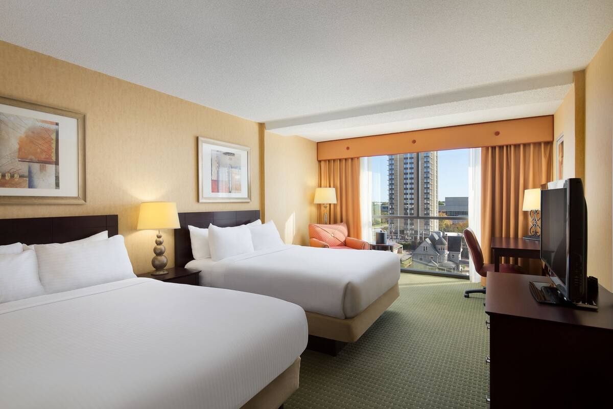 A hotel room with two beds, a desk with a TV, and a view of a cityscape.