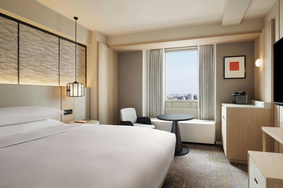 A hotel room with a large bed, a small round table, a chair, a desk, a television, and a window with curtains.