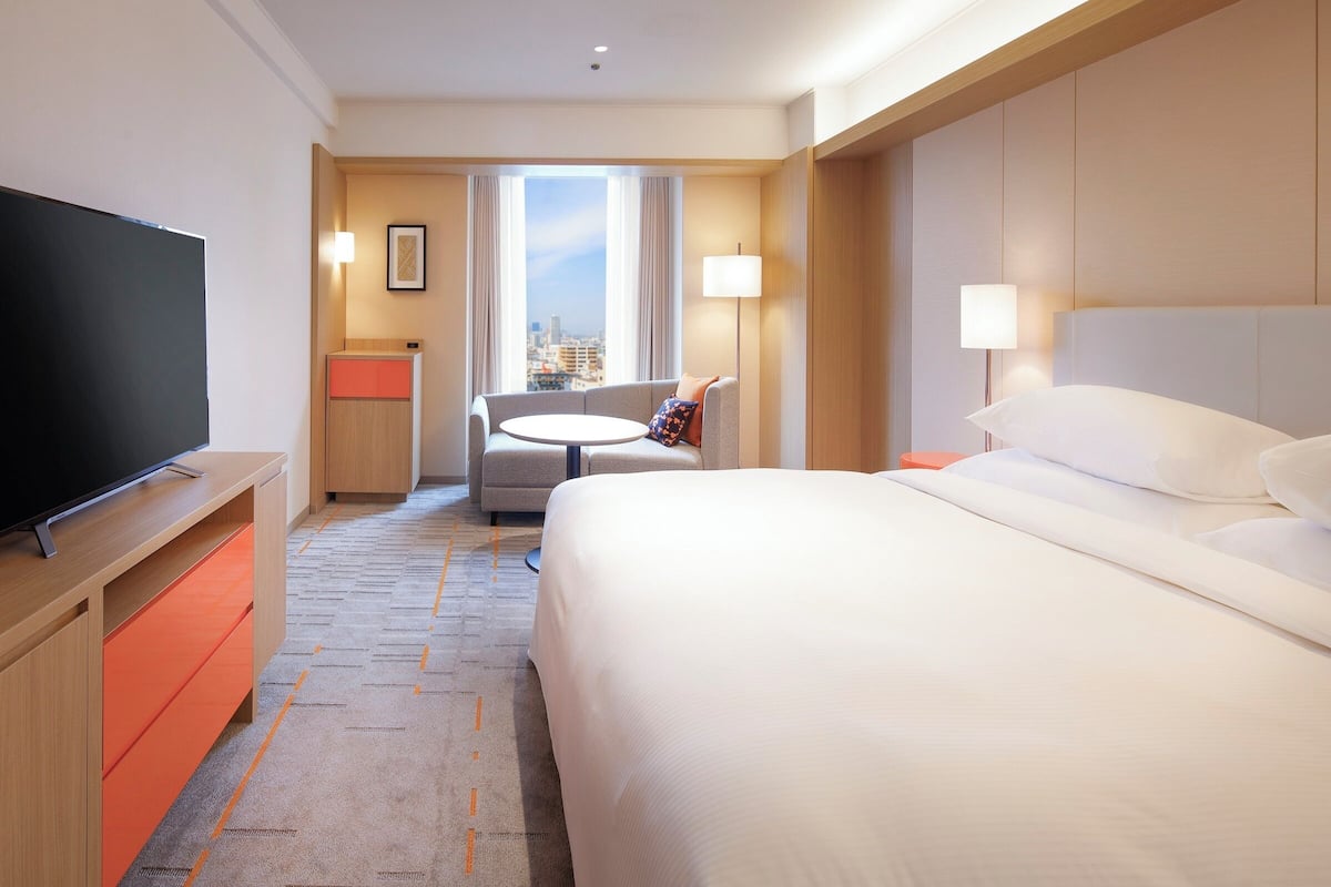 A hotel room with a bed, a TV, a desk, a chair, and a view of the city.