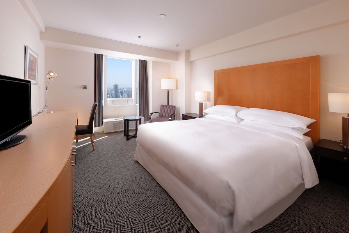 A hotel room with a large bed, a desk with a flat-screen TV, a chair, a nightstand with a lamp, and a window with a city view.