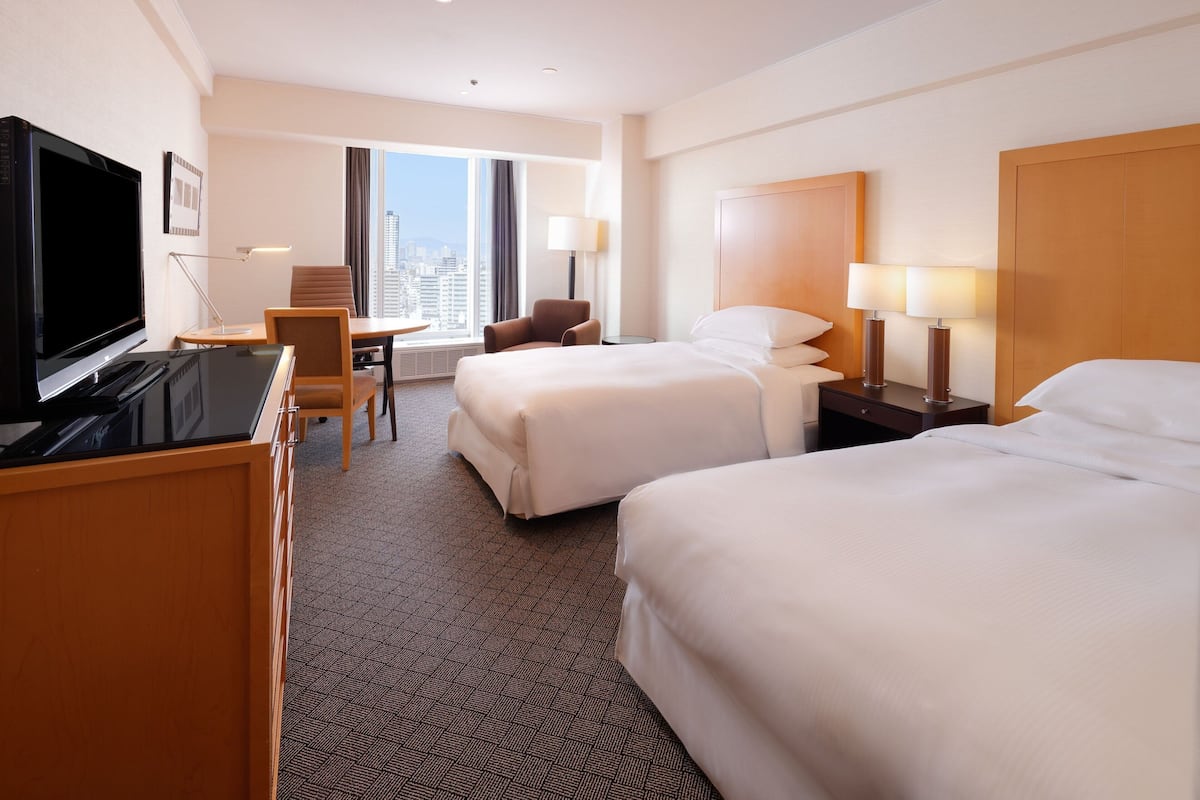 A hotel room with two beds, a desk with a TV, a chair, and a view of the city.
