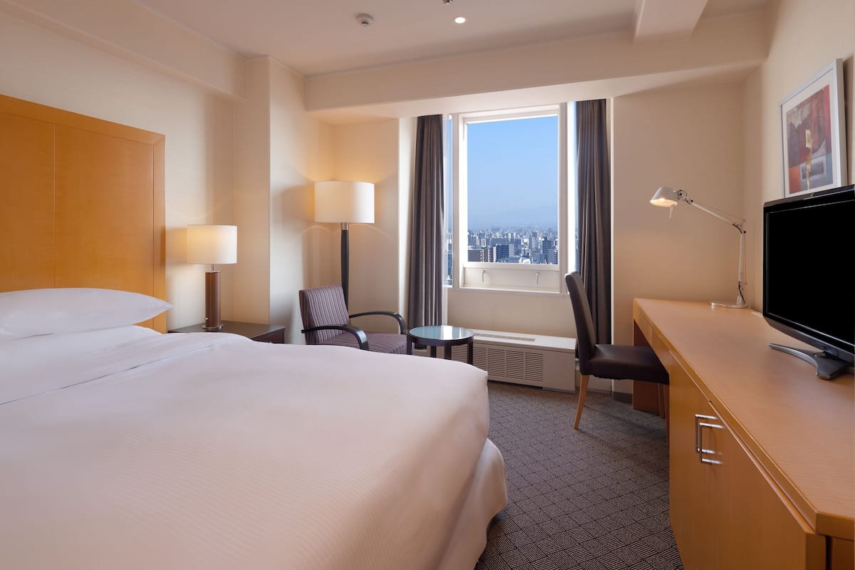 A hotel room with a large bed, a desk with a TV, a chair, a lamp, and a view of the city through the window.