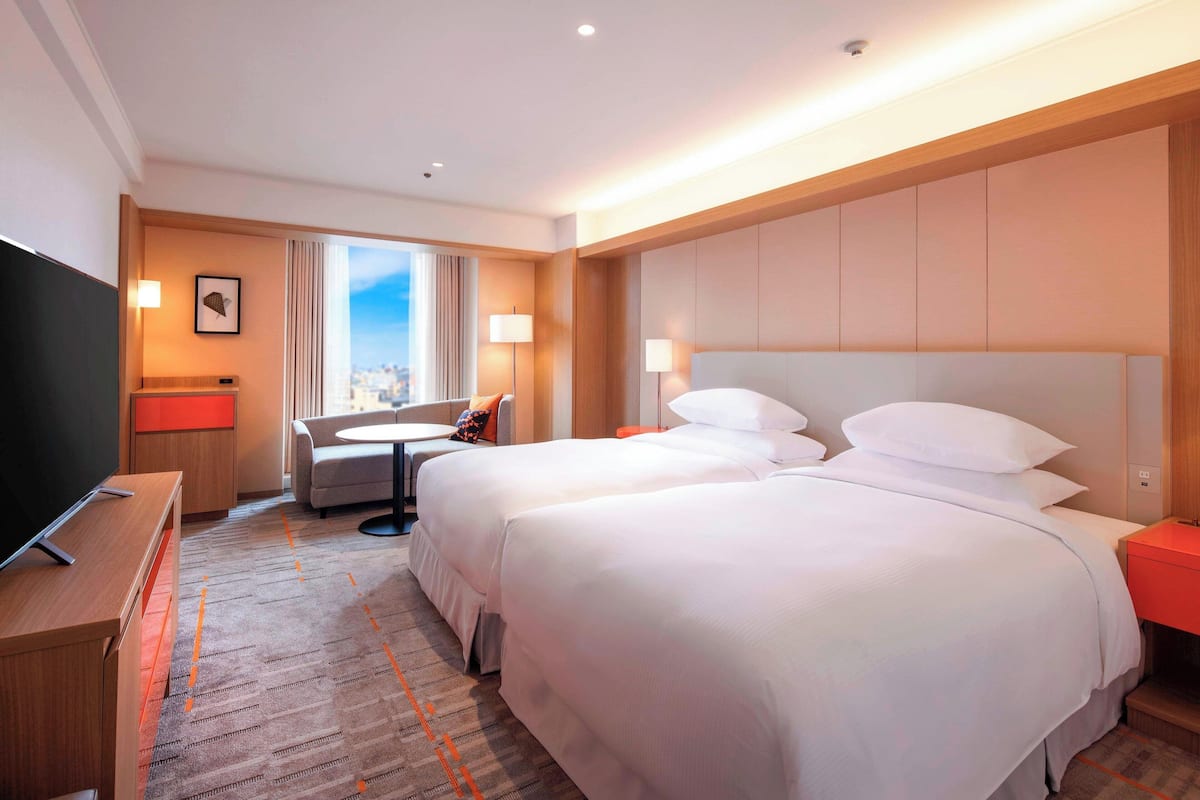A hotel room with two beds, a desk, a TV, and a city view.