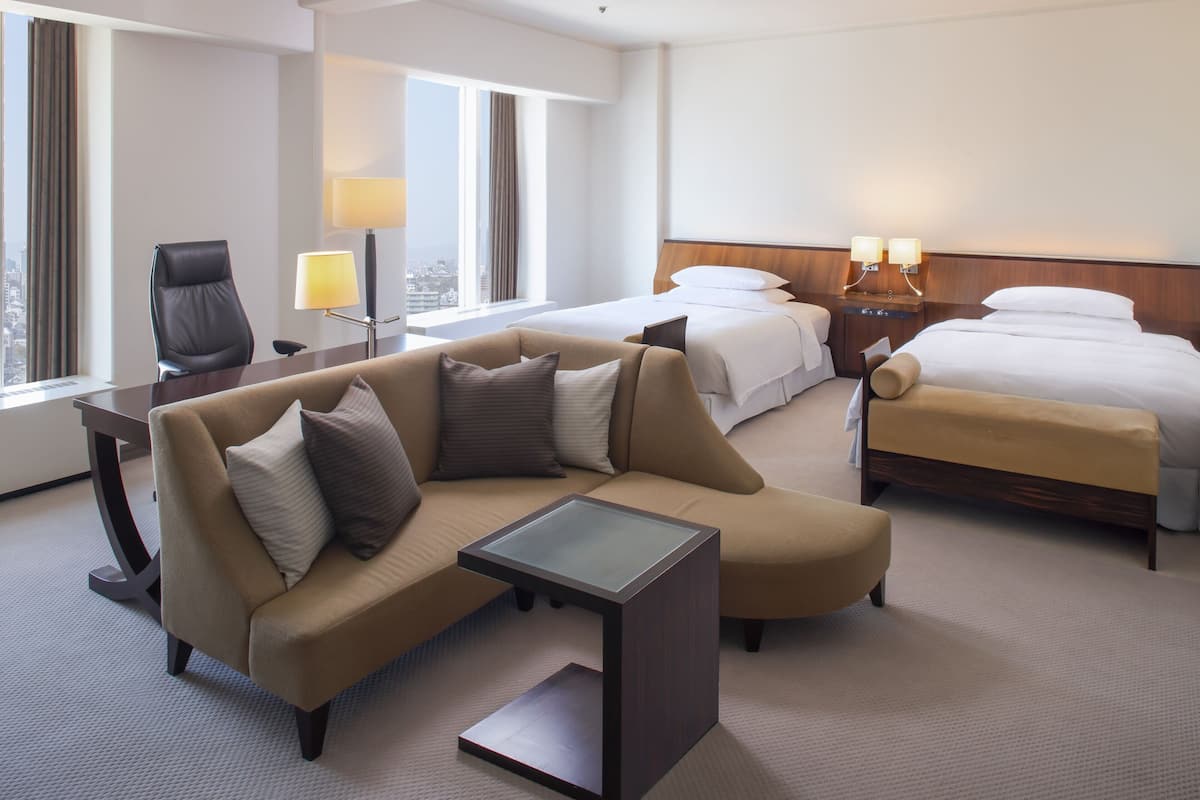 A hotel room with a sofa, two beds, a desk, and a lamp.