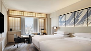 Habitación de hotel con dos camas, una zona de estar con una mesa, vistas a la ciudad y una obra de arte grande en la pared.