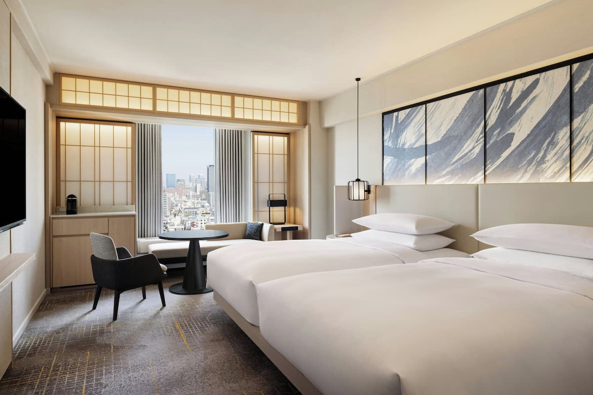 A hotel room with two beds, a seating area with a table, a city view, and a large artwork on the wall.