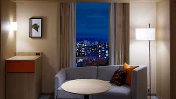 A hotel room with a sofa, a small table, a bedside table, a lamp, a framed picture, and a view of a cityscape at night.