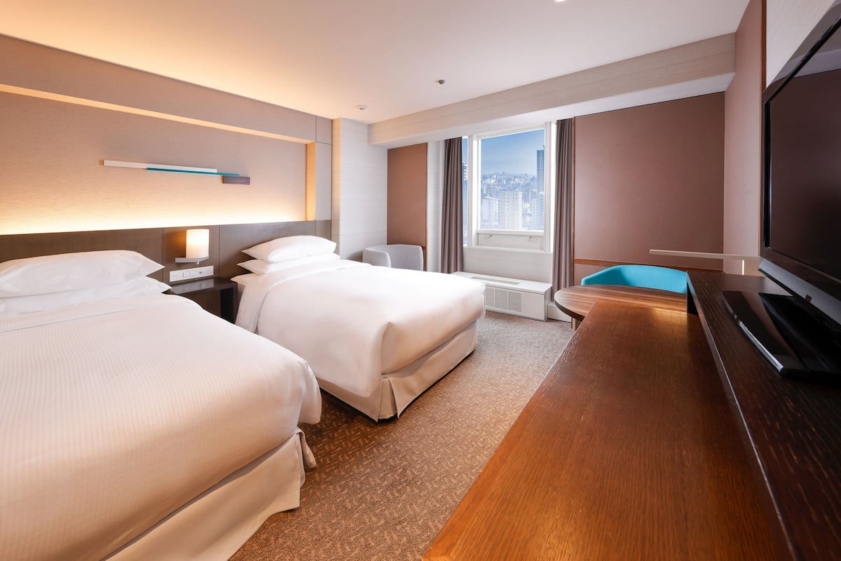 A hotel room with two beds, a TV, a desk, and a window with a city view.