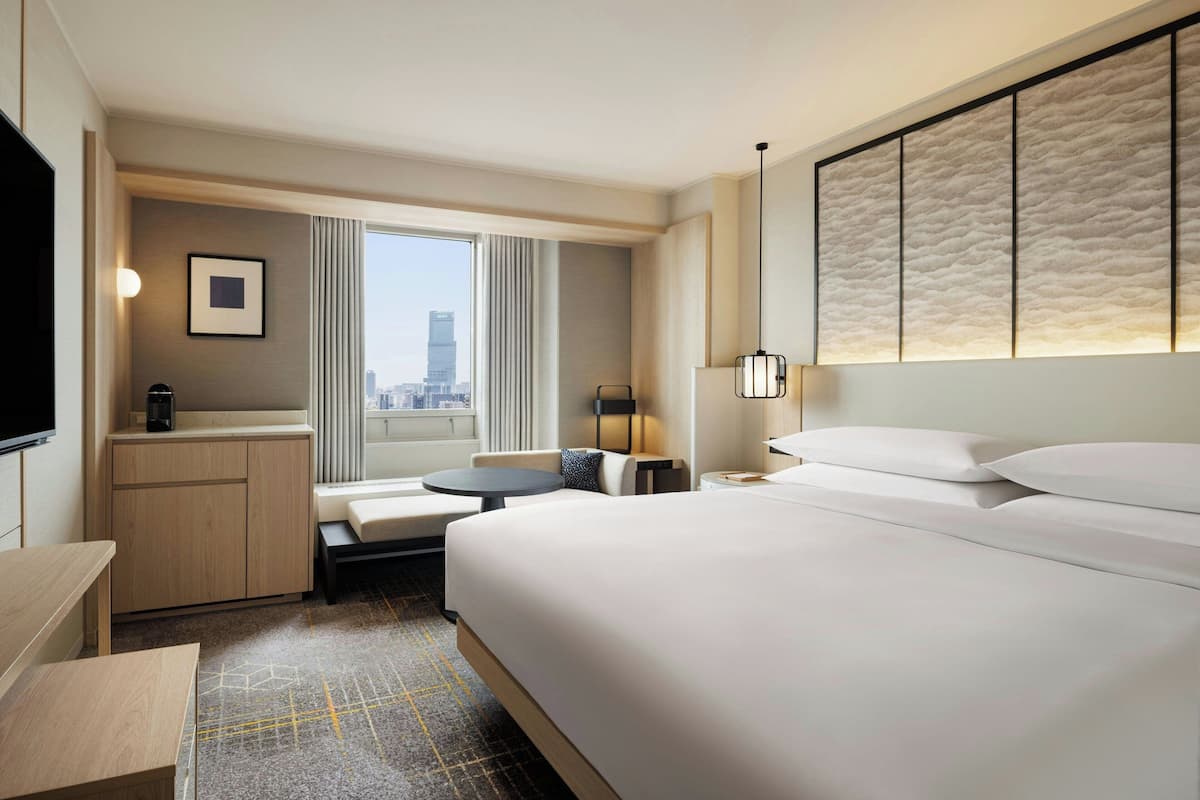 A hotel room with a large bed, a desk, a chair, a small table, a TV, and a window with a city view.