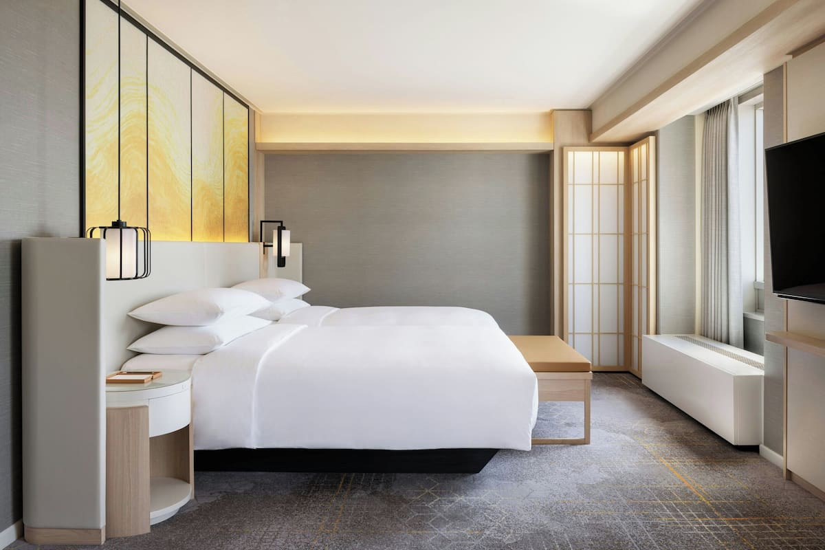 A modern hotel room with a large bed, bedside tables, a TV, and a view of the city through large windows.