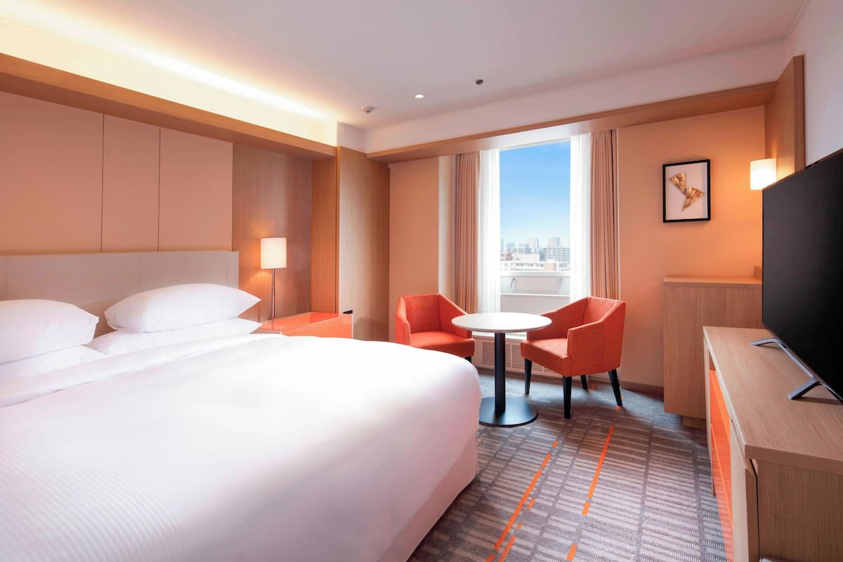 A hotel room with a large bed, two orange chairs, a small table, a TV, and a city view.