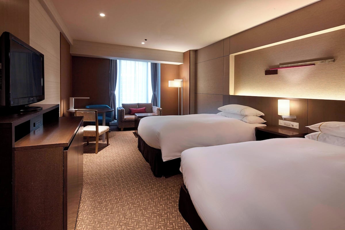A hotel room with two beds, a desk with a flat-screen TV, a chair, and a lamp.