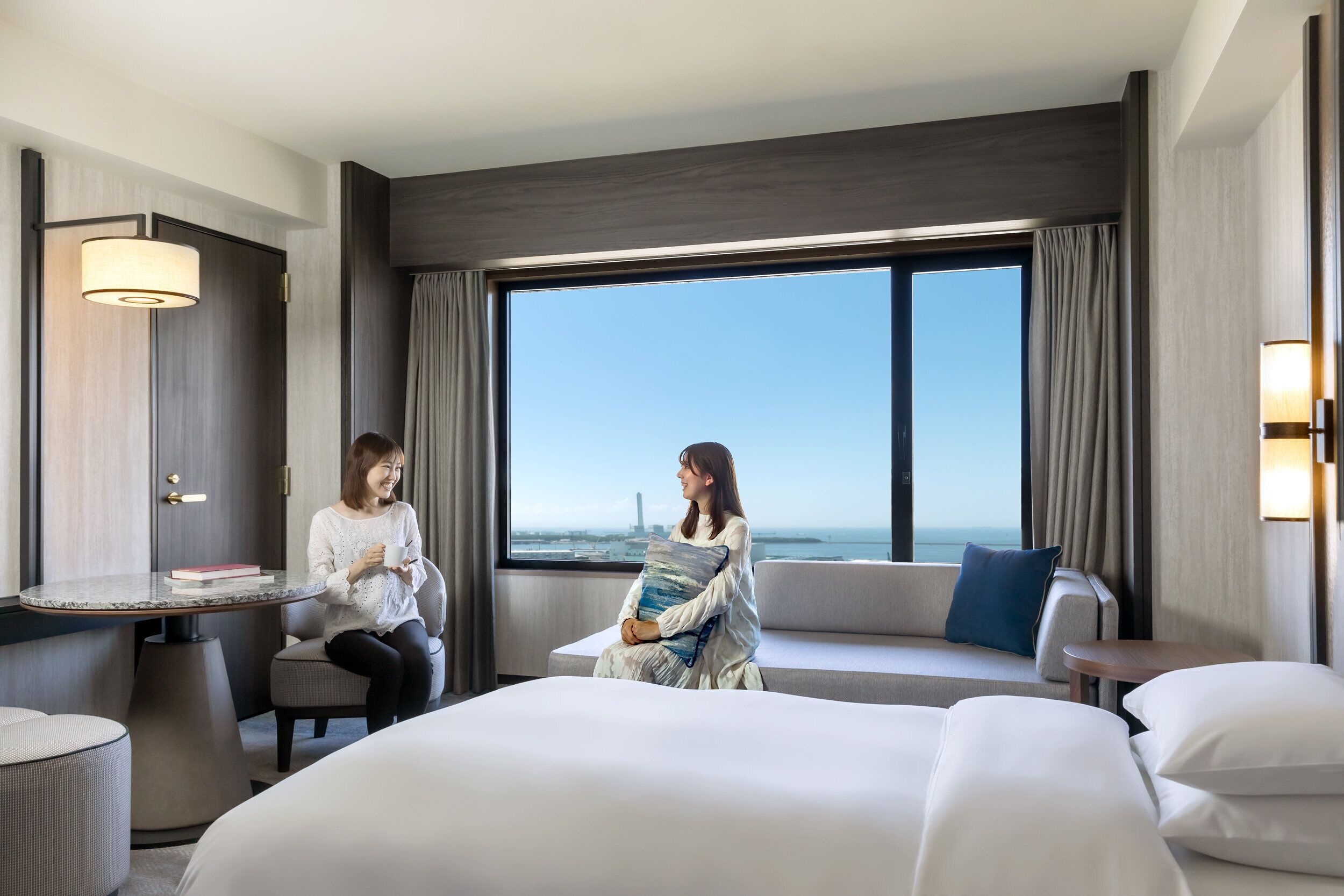 A hotel room with a large bed, a sofa, a small table, and a view of the cityscape through the window.