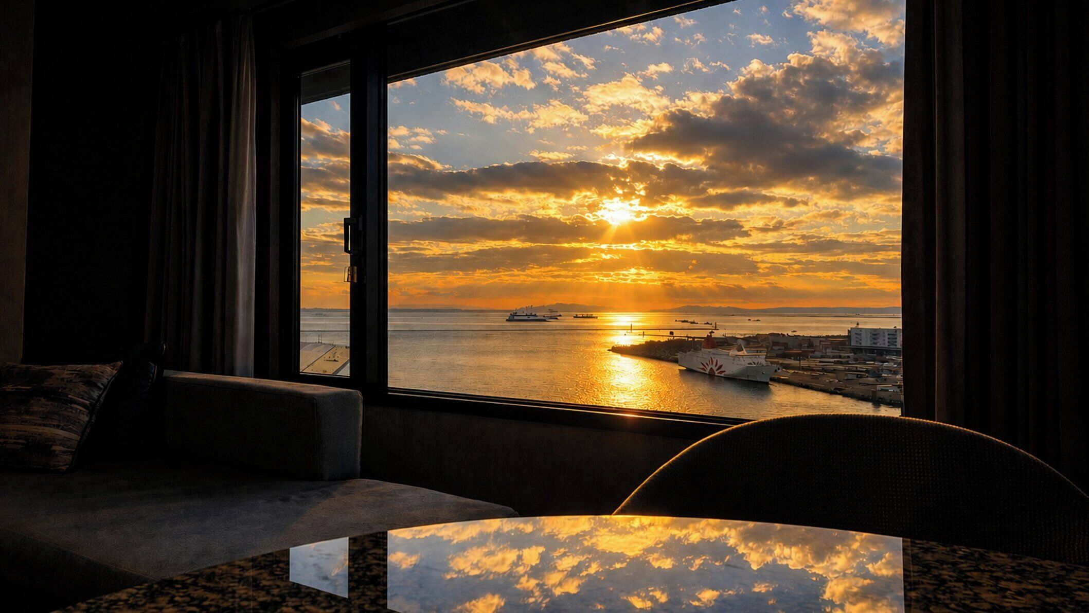 A room with a large window overlooking a sunset over a body of water, a sofa, and a small table.