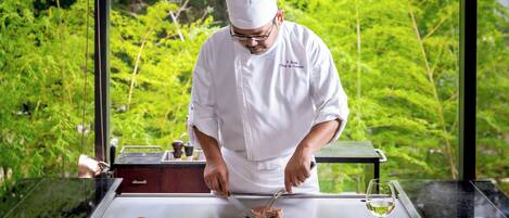 A chef preparing food at an outdoor kitchen with a view of lush greenery.