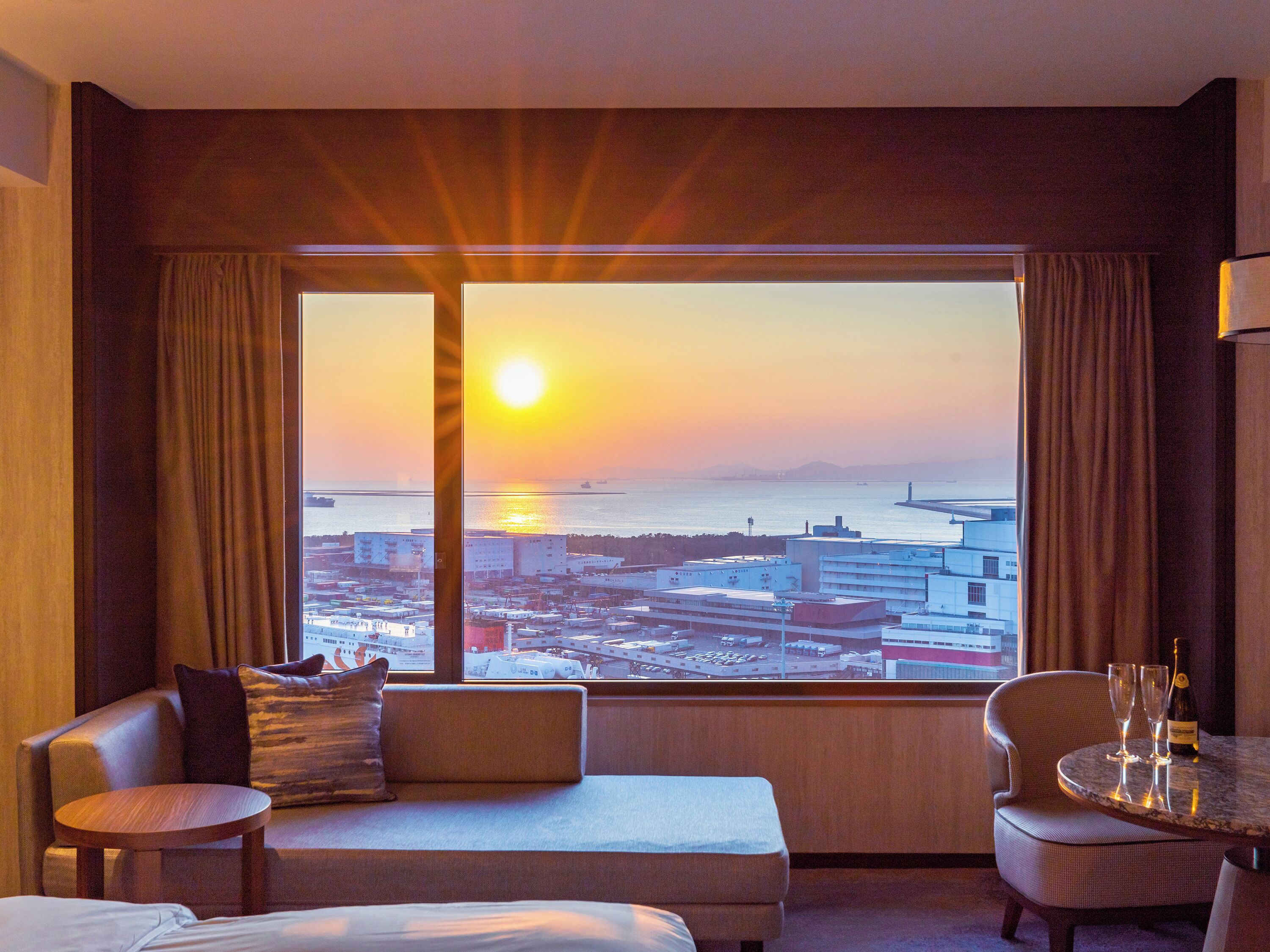 A hotel room with a large window, a sofa, a chair, and a small table with glasses and a bottle.